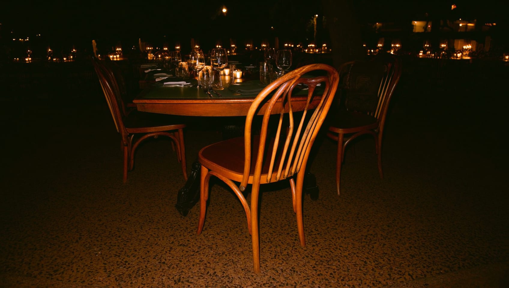 A single empty wooden dining chair pulled out from a dark restaurant table at night, warm candlelight beyond