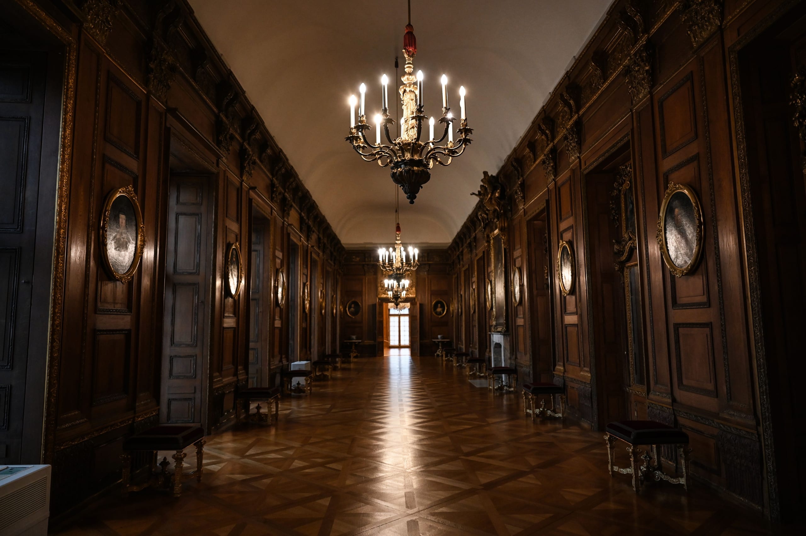 A dim wood-paneled corridor lined with framed portraits and hanging candle-style chandeliers receding into shadow