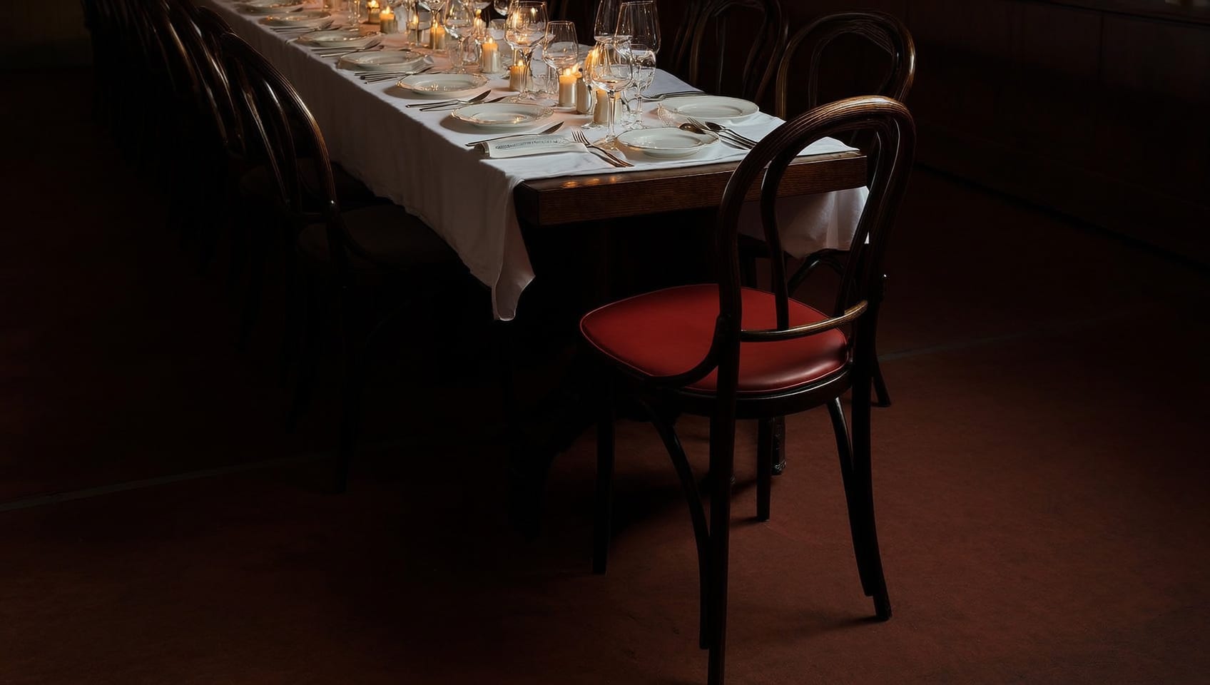 A long candlelit dinner table set for many, one dark chair pulled out and waiting in the foreground