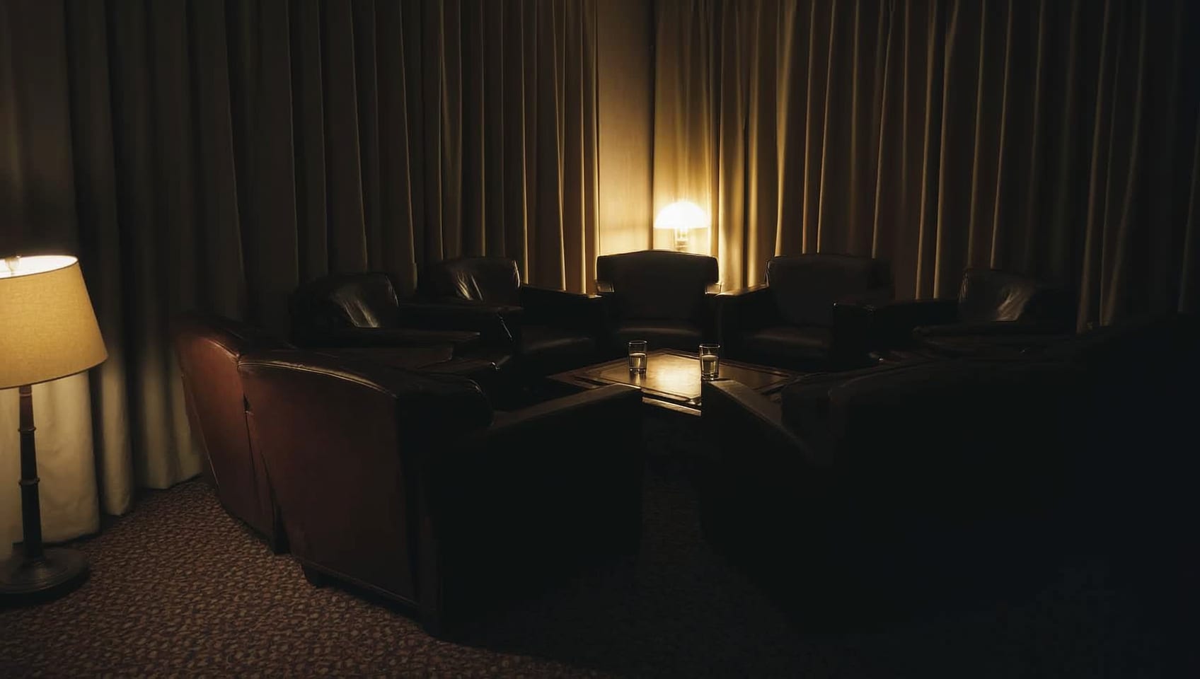 A ring of dark leather armchairs facing each other in a dim, curtained room, lit by a single lamp