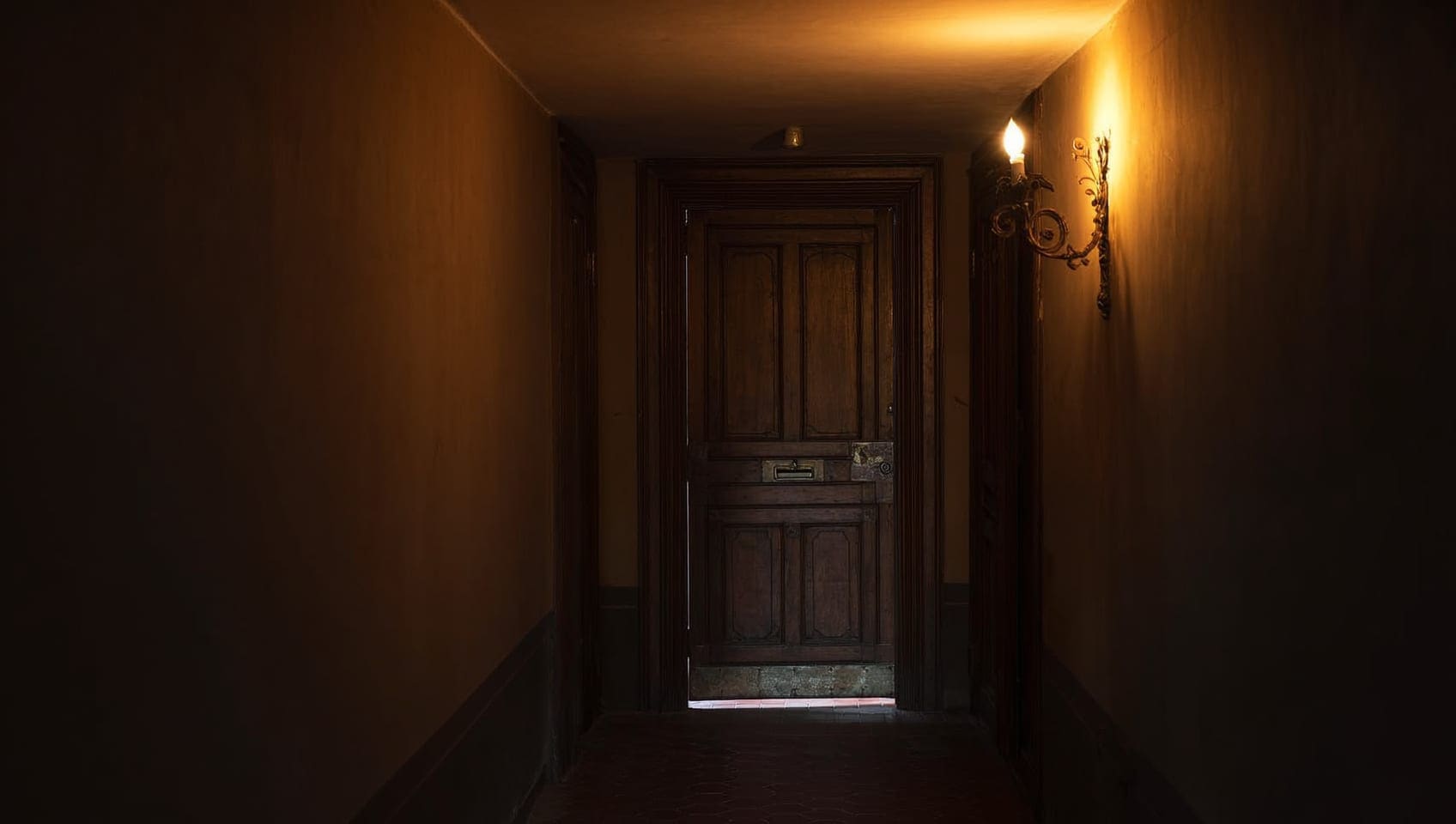 A dim wood-paneled corridor leading to a closed unmarked door lit by a single wall sconce